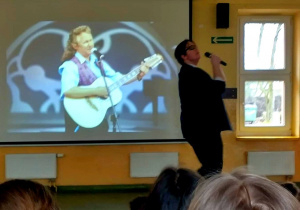 Chłopak stoi na scenie w okularach przeciwsłonecznych, w ręku trzyma mikrofon; improwizuje śpiew; za nim na ekranie widać oryginalne wideo z nagraniem.