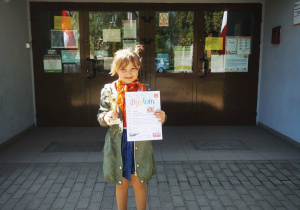 Uczennica reprezentująca szkołę w konkursie wiedzy o Łasku pozuje do zdjęcia prezentując otrzymany dyplom oraz puchar. W tle widać budynek szkoły.