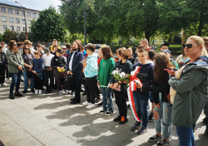 Uczniowie zgromadzeni przed pomnikiem ofiar Holocaustu słuchają w skupieniu przewodnika wycieczki.