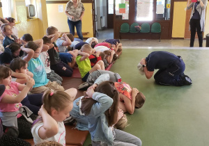Pani policjant prezentuje dzieciom zasady przechodzenia do pozycji "żółwia" w sytuacji ataku agresywnych psów. Dzieci naśladują.