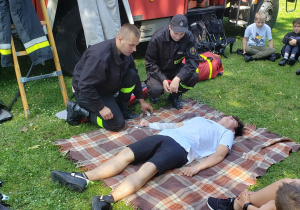 Strażacy demonstrują na uczniu sposób unieruchomienia kończyny w przypadku złamania.