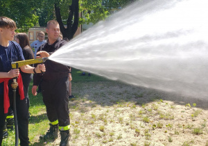 Uczeń leje wodę z węża strażackiego na boisku szkolnym.