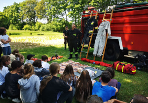 Uczniowie siedzą na trawie, słuchając z zainteresowaniem strażaka, który instruuje jak udzielać pierwszej pomocy.