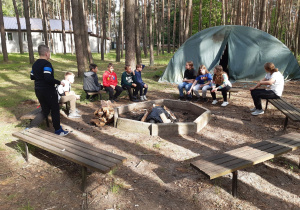 Uczniowie siedzą w lesie wokół miejsca na ognisko. W tle widać namiot wojskowy.