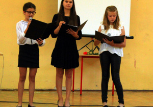 Trzy uczennice odczytują do mikrofonów swoje kwestie podczas części artystycznej na zakończenie roku szkolnego.