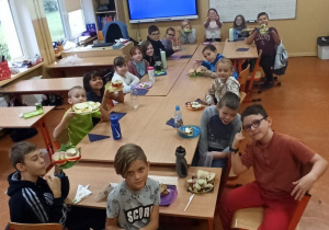 Uczniowie klasy trzeciej siedzą przy dużym stole w klasie, prezentując własnoręcznie wykonane kanapki.
