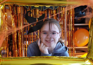 Uczennica w ramce na tle dekoracji Halloweenowej. Przystawia do głowy kapelusz czarownicy na patyku.