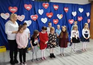 Uczniowie grupy przedszkolnej podczas występu dla babć i dziadków. W tle dekoracja z sercami.
