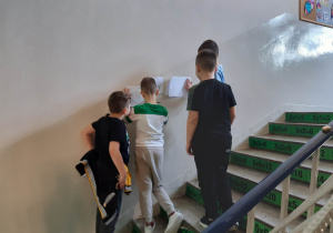 Uczniowie rozwiązują łamigłówki matematyczne przyczepione na ścianie korytarza szkolnego.