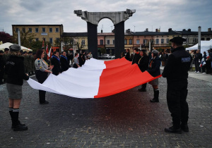 Harcerze z wielką biało-czerwoną flagą na tle pomnika na placu 11 Listopada w Łasku