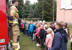 Grupa dzieci słucha z zainteresowaniem strażaków prezentujących wóz strażacki.