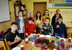 Uczniowie klasy czwartej pozują do zdjęcia przy stole zastawionym słodkościami i napojami przyniesionymi z okazji Dnia Chłopaka.