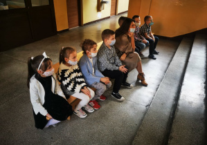 Uczniowie klasy pierwszej wraz z wychowawczynią siedzą na ławce na szkolnym korytarzu w oczekiwaniu na uroczysty apel z okazji Dnia Edukacji Narodowej.