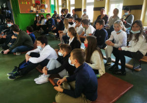 Uczniowie starszych klas wraz z nauczycielami siedzą na materacach i ławkach na szkolnym korytarzu przyglądając się przedstawieniu.