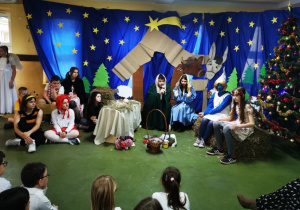 Przebrani uczniowie biorący udział w przedstawieniu bożonarodzeniowym siedzą na tle dekoracji przedstawiającej stajenkę.