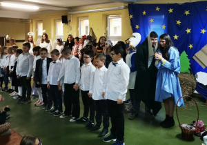 Uczniowie klasy trzeciej w odświętnych strojach śpiewają piosenkę bożonarodzeniową przed widownią szkolną.