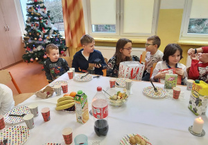 Uczniowie klasy piątej i pierwszej siedzą przy wigilijnym stole, pokazują sobie nawzajem otrzymane właśnie przezenty.