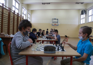 Dwóch chłopców rozgrywa partię szachową. Jeden z nich przestawia pionki na szachownicy. W tle widać innych graczy i stoliki z rozstawionymi na nich planszami i pionkami do gry.