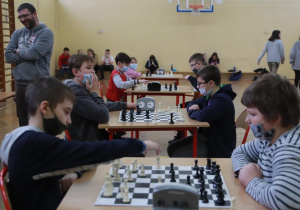 Dwóch chłopców rozgrywa partię szachową. Jedna z nich przestawia pionki na szachownicy. W tle widać innych graczy i stoliki z rozstawionymi na nich planszami i pionkami do gry.